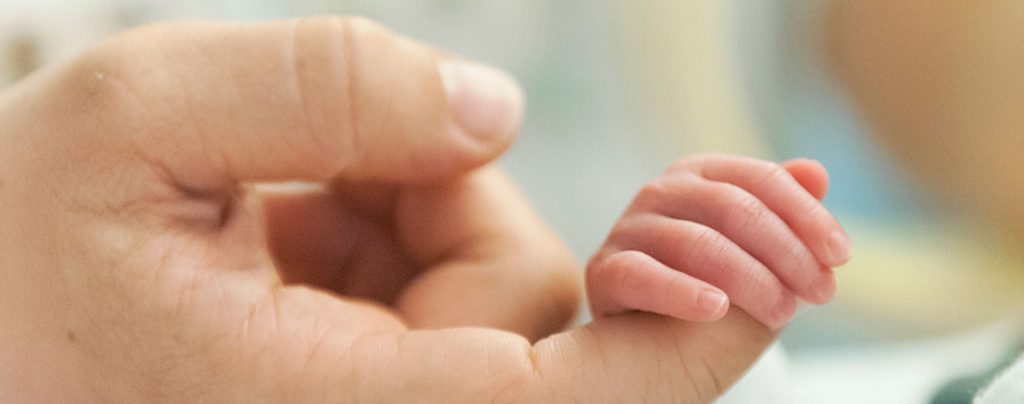 La main d'un tout jeune nourrisson en néonatalogie tenue par celle d'un adulte.